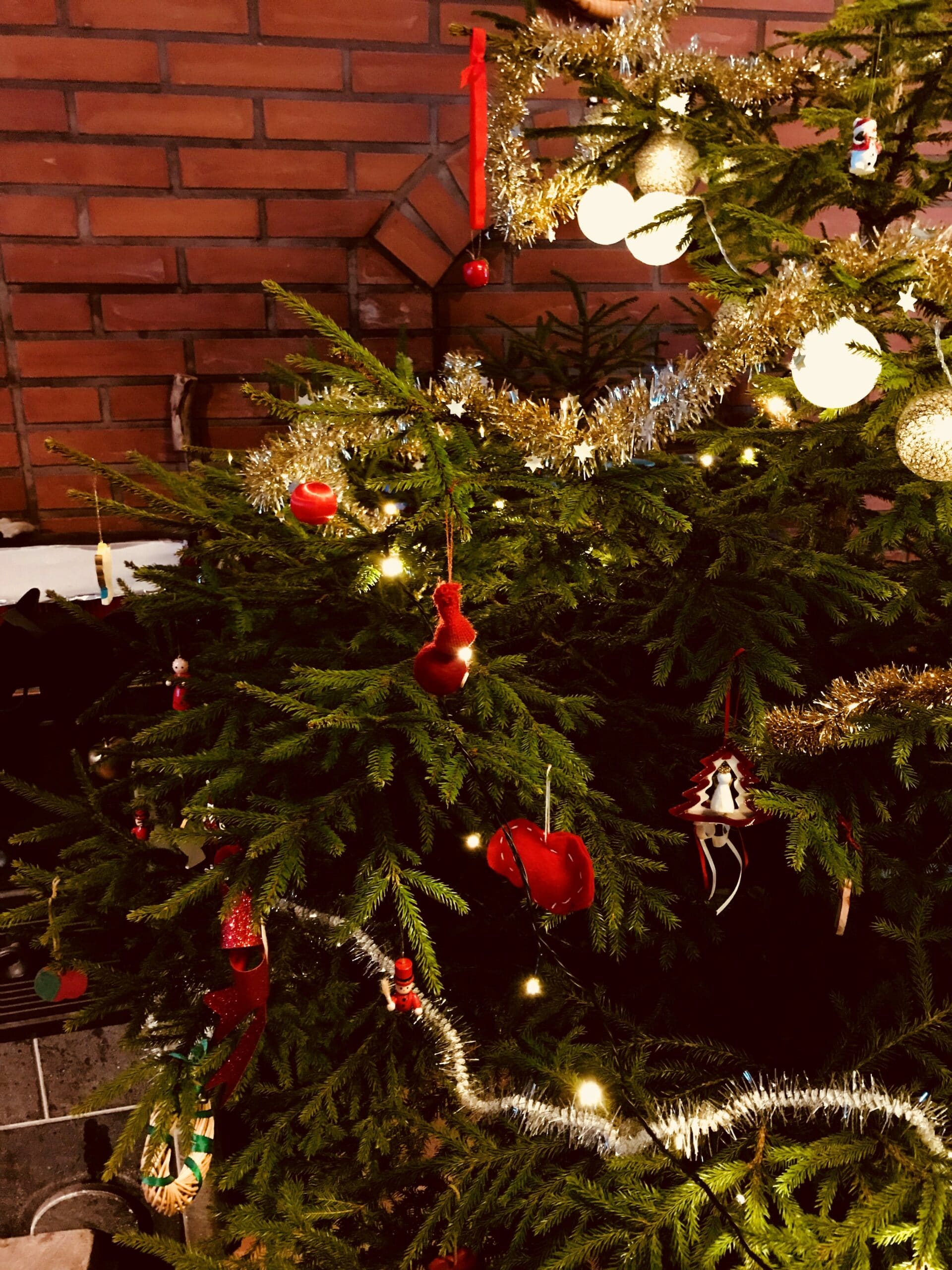 Kuusi, jossa on punaisia ja valkoisia joulukoristeita. Kuusen takana näkyy punatiilinen takka.