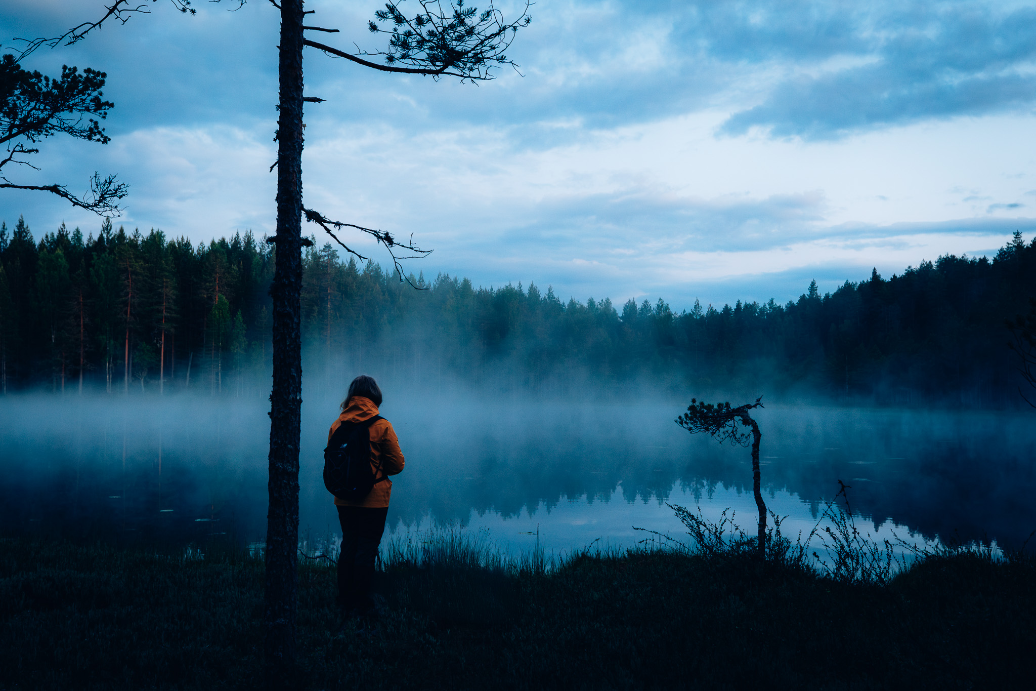 Oranssiin takkiin pukeutunut henkilö seisoo veden rannalla. Vedestä nousee usvaa. On hämärä.
