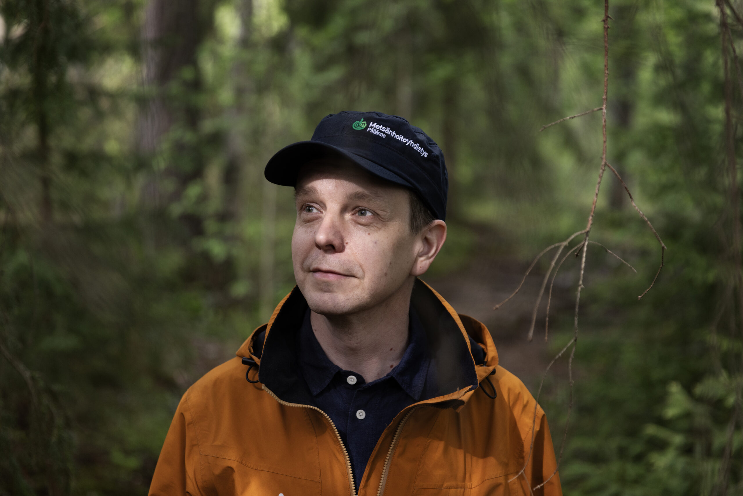 Mies oranssissa takissa ja lippalakissa katsoo hieman ohi kameran. Taustalla puita ja lehvästöä.
